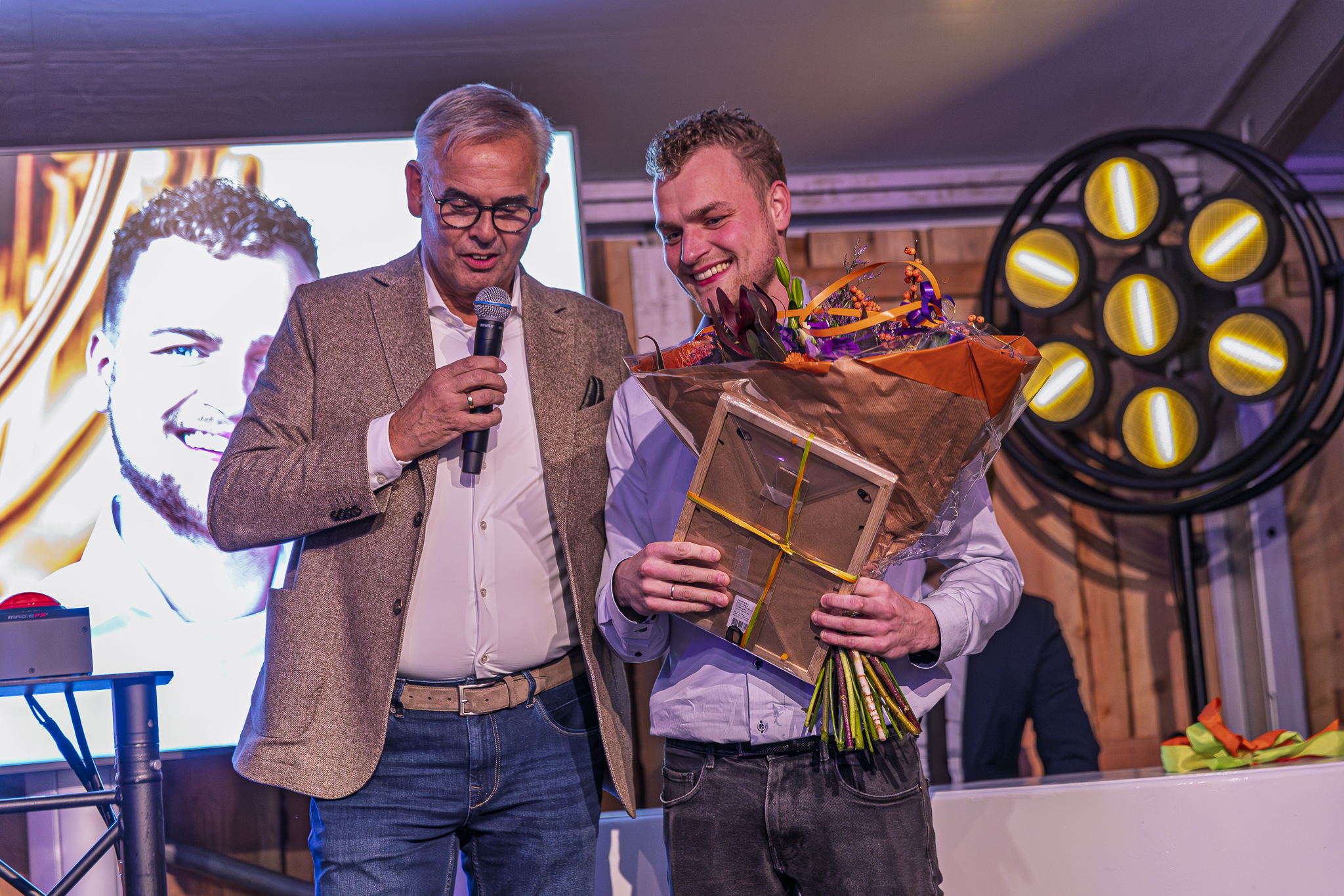 Speechende man met microfoon naast een lachende jongere man met een bos bloemen en lijst in zijn handen - Timeless Events
